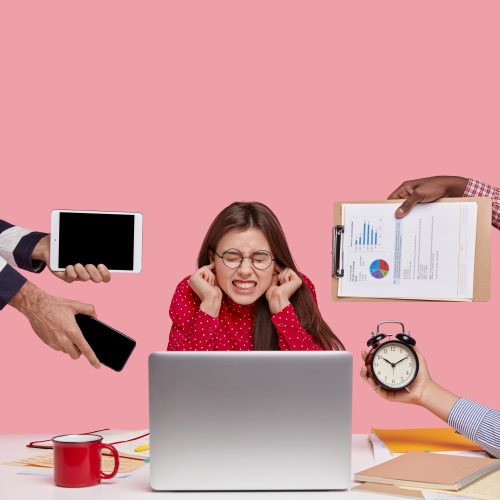 Vertical shot of dissatisfied woman works in office, covers ears, ignores reproaches of colleagues who bother her on different questions, works on modern laptop computer, isolated on pink background