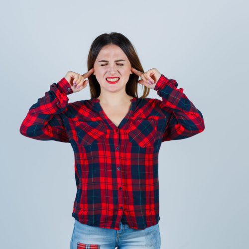 young woman plugging ears with index fingers, keeping eyes closed in checked shirt, jeans and looking harried , front view.